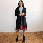 Woman wearing a knee length brick colour A-line skirt with a light grey turtleneck top and a long black jacket