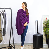 Woman wearing a purple poncho standing next to a black suitcase in a room with a clothing rack and plant.