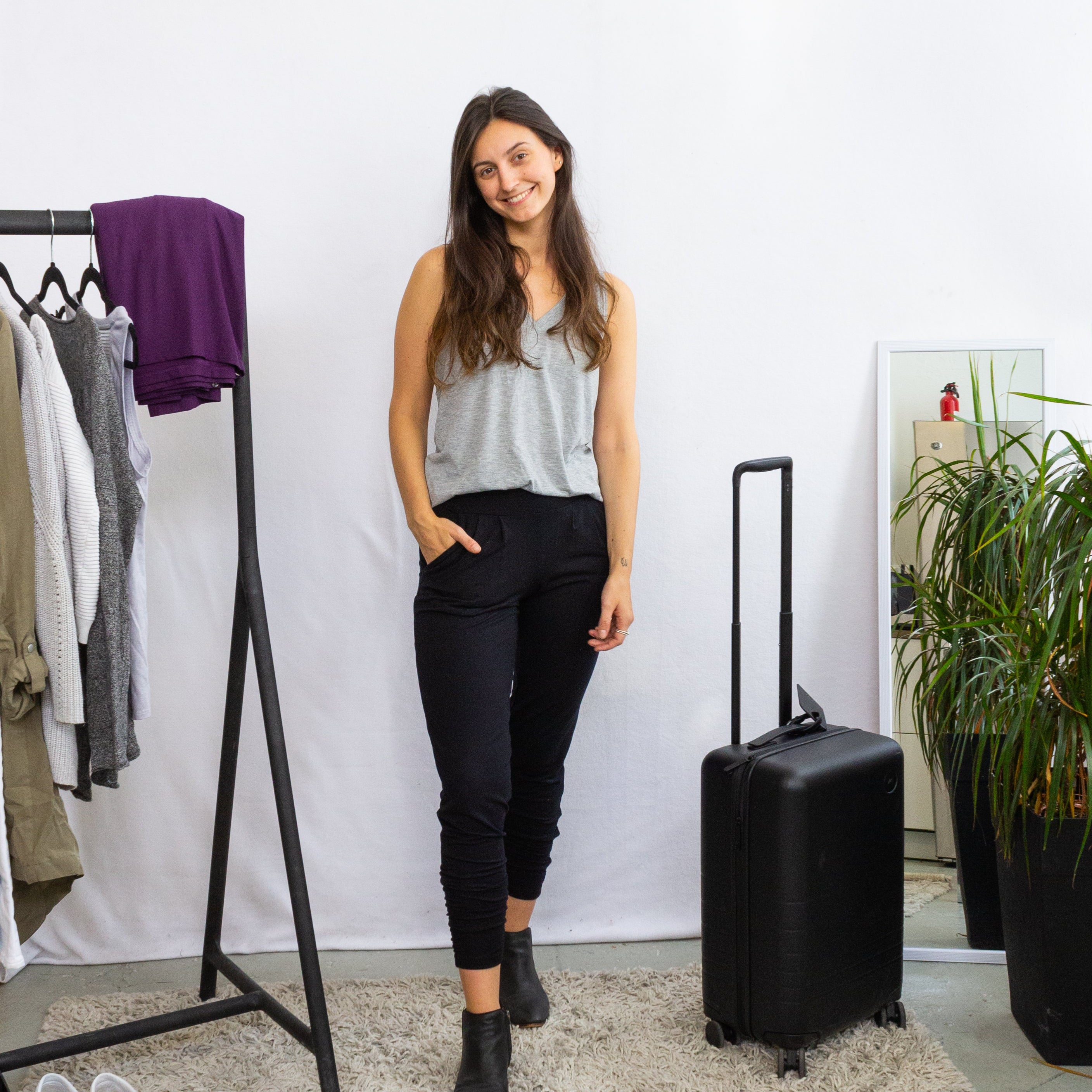 Woman standing next to a black suitcase in a room with a clothing rack and plants.