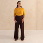 woman wearing a yellow boat neck blouse and aubergine trouser wide leg pants with brown shows and golden earrings