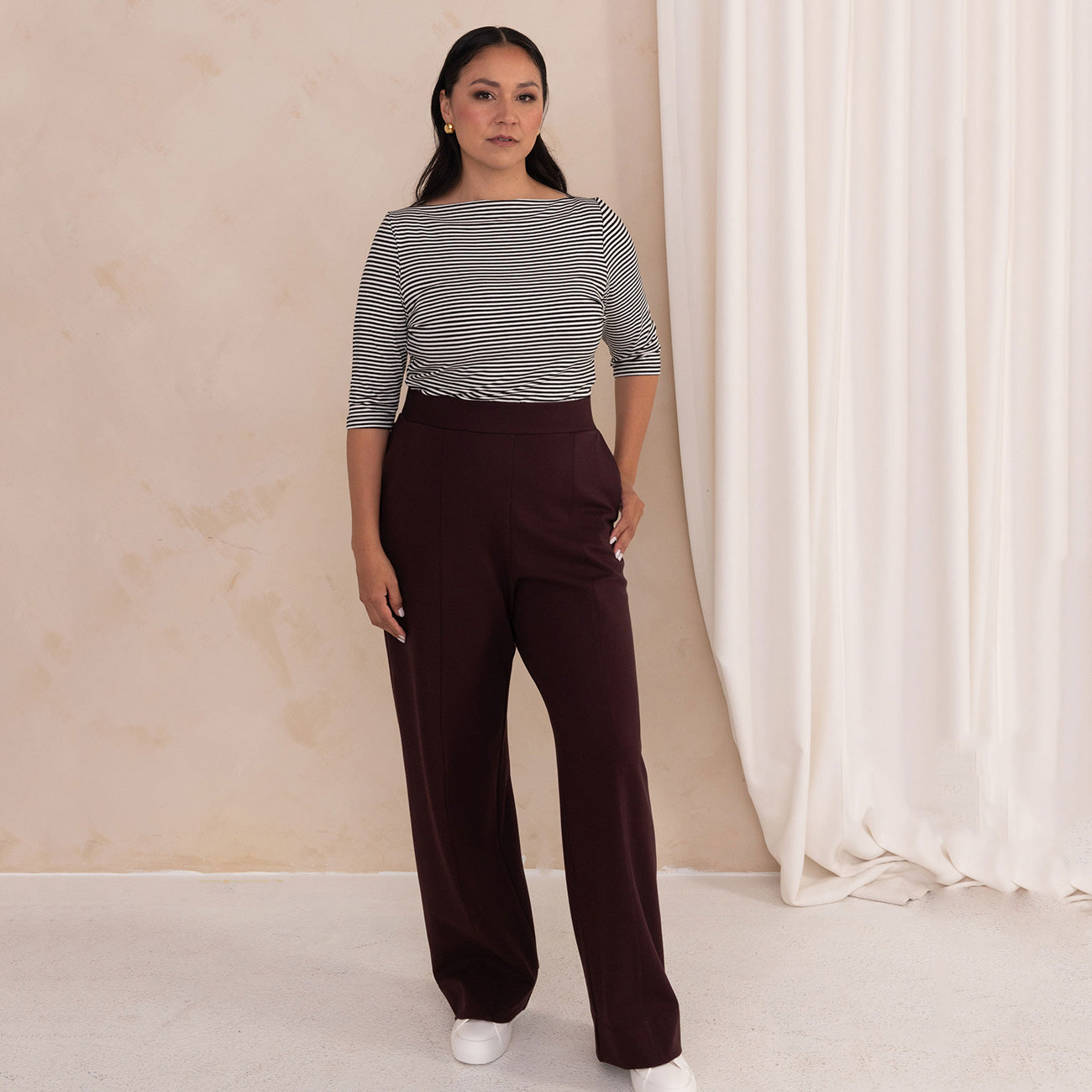 woman wearing a black and white stripes boat neck top  and high waisted aubergine wide leg trouser pants with white sneakers