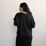 Woman showing the back and wearing a grey turtleneck longlseeve blouse with a black v-neck sweater layered on top