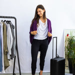 Woman in a stylish outfit with a purple cardigan, white shirt, and black pants, standing next to a black suitcase in a room with a rack of clothes and shoes.