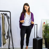 Woman in a stylish outfit with a purple cardigan, white shirt, and black pants, standing next to a black suitcase in a room with a rack of clothes and shoes.