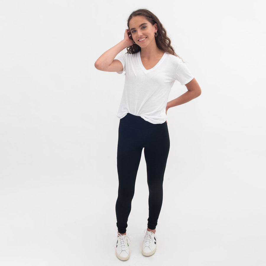 Woman wearing tight fitted black stretchy leggings with horizontal ribbing on knees featuring zippers on the back pockets and ankle and white v-neckline t-shirt