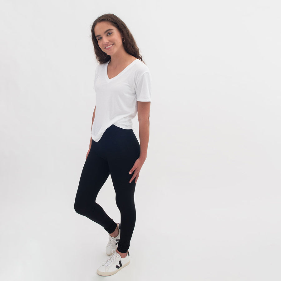 Woman wearing tight fitted black stretchy leggings with horizontal ribbing on knees featuring zippers on the back pockets and ankle and white v-neckline t-shirt