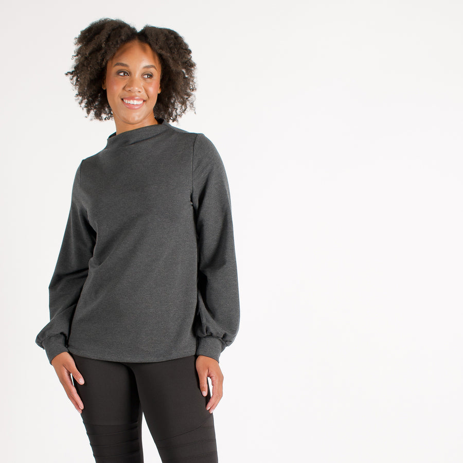 Woman wearing dark grey loose blouse with puffy long sleeve and funnel neckline with black fitted leggings