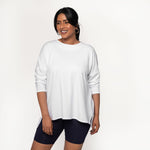 Woman wearing white long sleeve shirt with a long length relaxed fit, front scoop neckline or v-neckline with pleating and side slits paired with tight black bike shorts