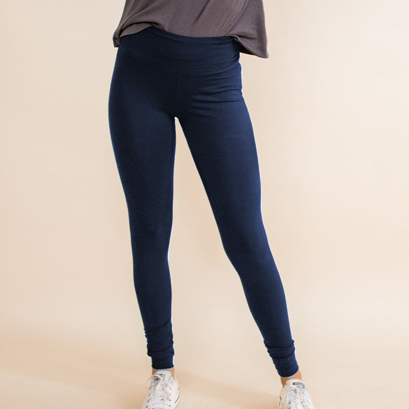 Woman wearing tight fitted dark blue stretchy leggings with horizontal ribbing on knees featuring zippers on the back pockets and ankle and grey long sleeve shirt