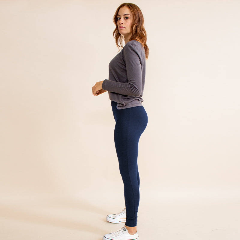 Woman wearing tight fitted dark blue stretchy leggings with horizontal ribbing on knees featuring zippers on the back pockets and ankle and grey long sleeve shirt