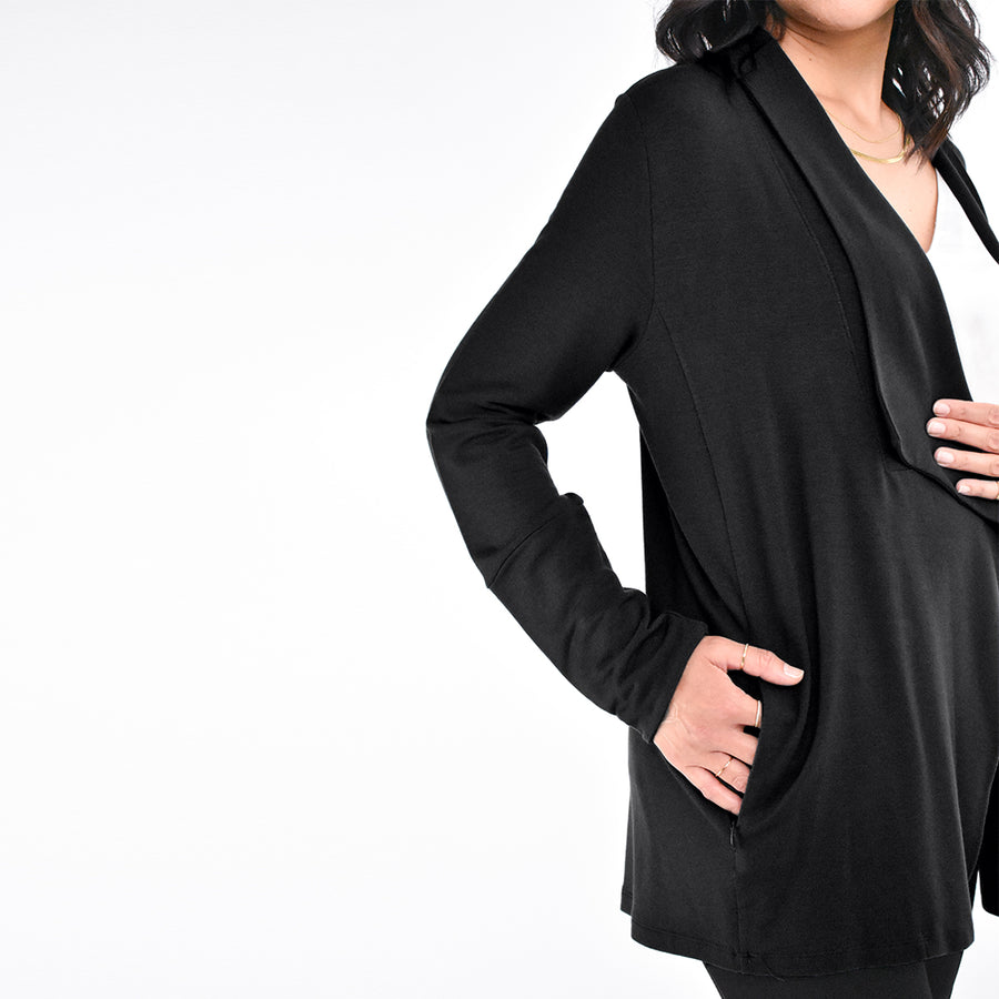 Woman wearing an open loose flowing black cardigan with wide flowing lapel and pockets with a white v-neck shirt and black leggings